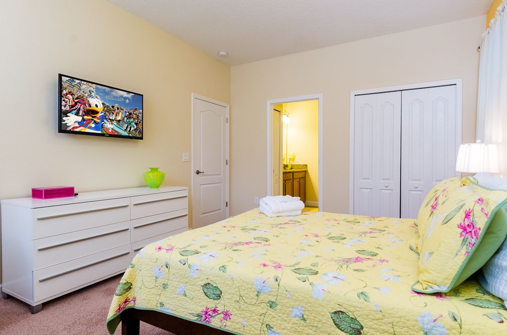 Un dormitorio con edredón estampado de flores, un aparador con cajones, un televisor en la pared y un armario con puertas.
