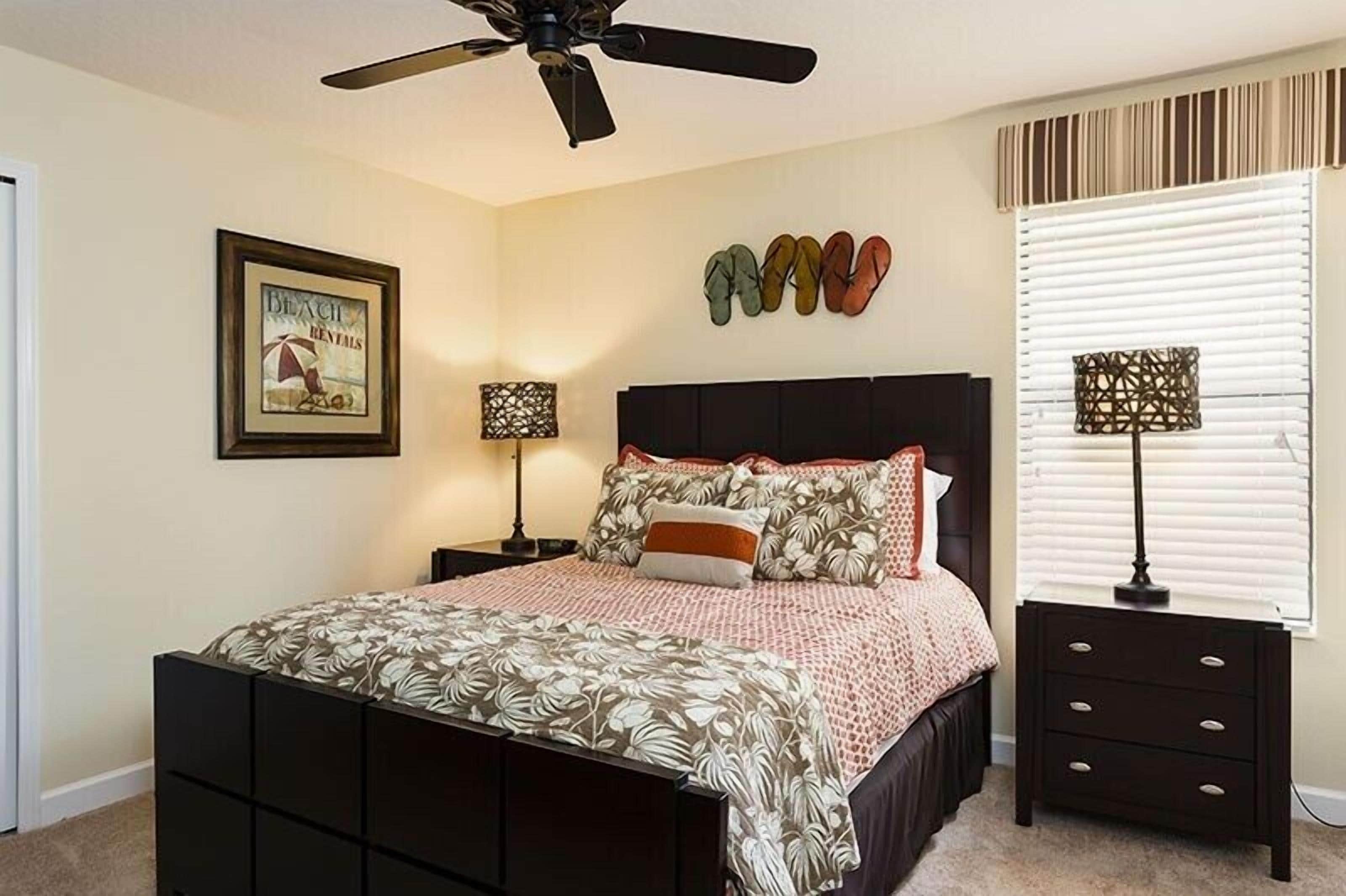 Un dormitorio con cama, ventilador de techo, cuadro enmarcado, lámpara de pared y ventana con persianas.