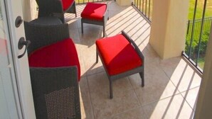Un balcon avec deux fauteuils en osier et des coussins rouges, un ventilateur de plafond et une vue sur une maison et un paysage verdoyant.