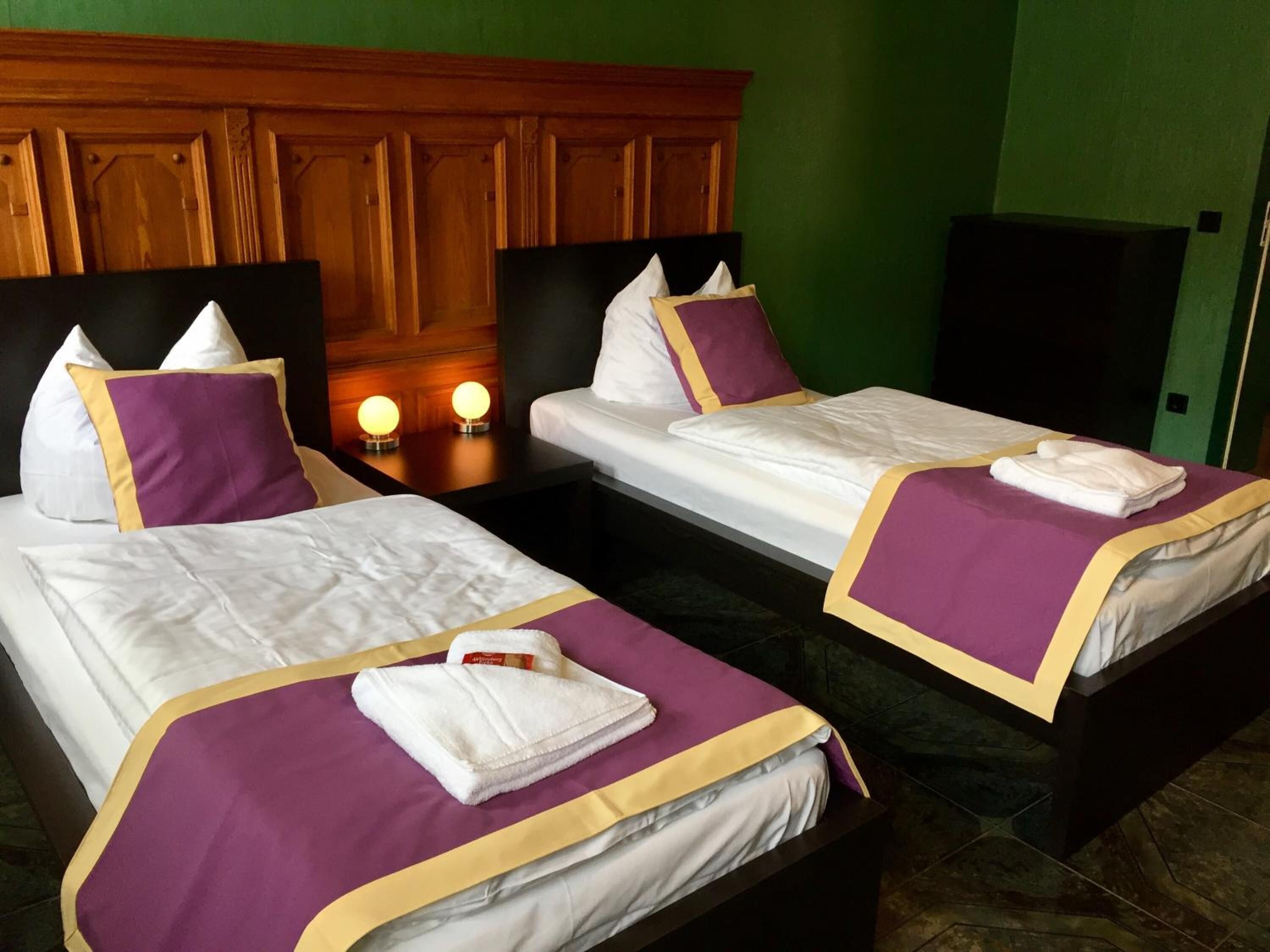 Ein Hotelzimmer mit zwei Betten, jeweils mit weißen Laken und violetten Akzenten. Auf den Nachttischen stehen Nachttischlampen und an der Wand ist ein Fernseher angebracht.