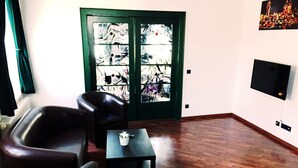 Een kamer met een houten vloer, twee zwarte leren stoelen, een tafeltje met een plant, een deur met een ruit en een tv aan de muur.