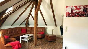 Een loft-stijl kamer met een bruine leren zithoek, een witte salontafel en een klein poufje. De kamer heeft zichtbare houten balken, een radiator en een schilderij aan de muur.