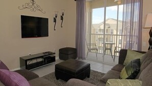 A living room with a sofa, ottoman, TV, and a balcony with a table and chairs. - Ov2939 - Windsor Hills Resort - 3 Bed 2 Baths Townhome (Kissimmee)