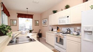 A modern kitchen with white cabinets, a stainless steel sink, a microwave, an oven, and a refrigerator. There are two windows with curtains, a hanging light fixture, and a few potted plants. - Ov2929 - Windsor Hills Resort - 5 Bed 5 Baths Villa (Kissimmee)