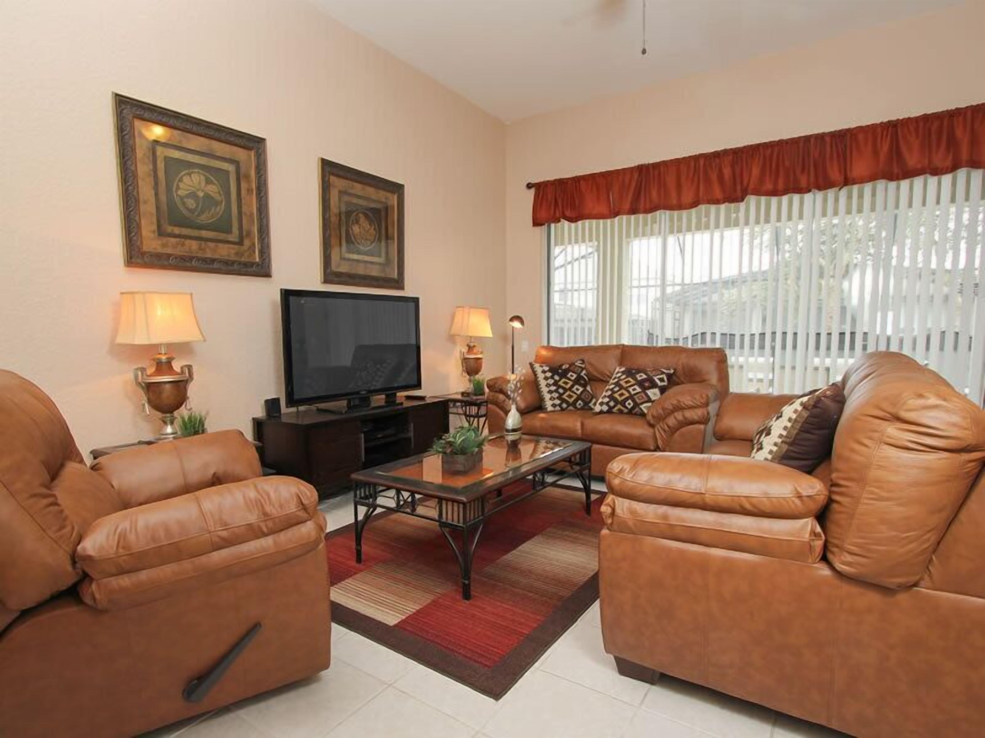 A living room with leather sofas, a flat-screen TV, two wall-mounted lamps, and a large window with blinds.