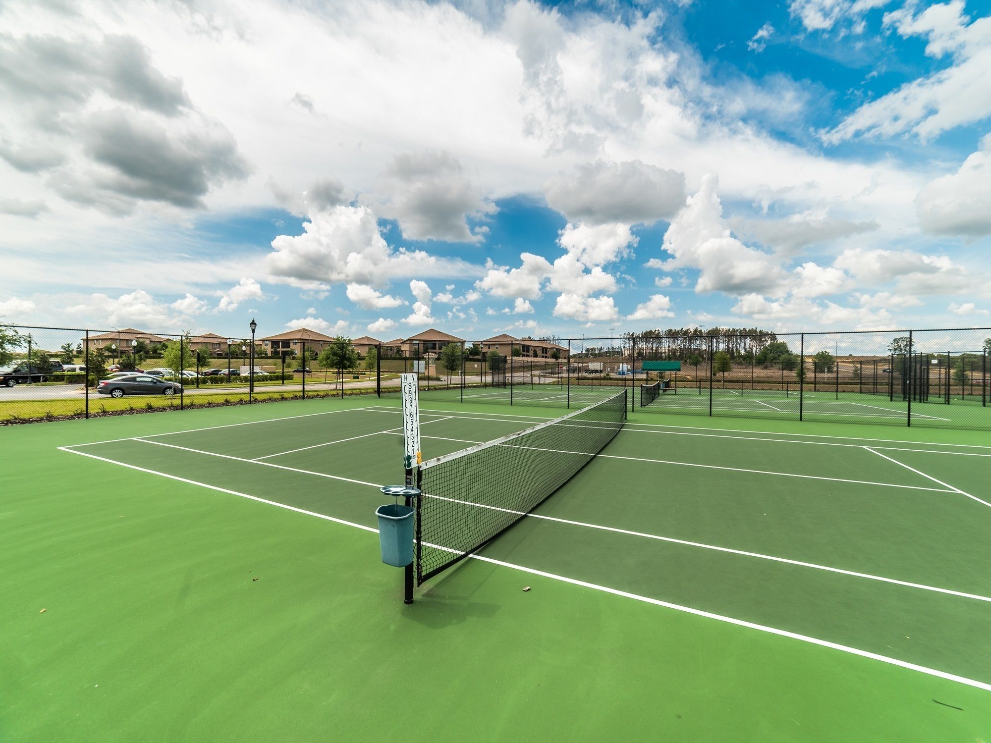 A tennis court with a net, surrounded by a fence, and a parking area with cars.