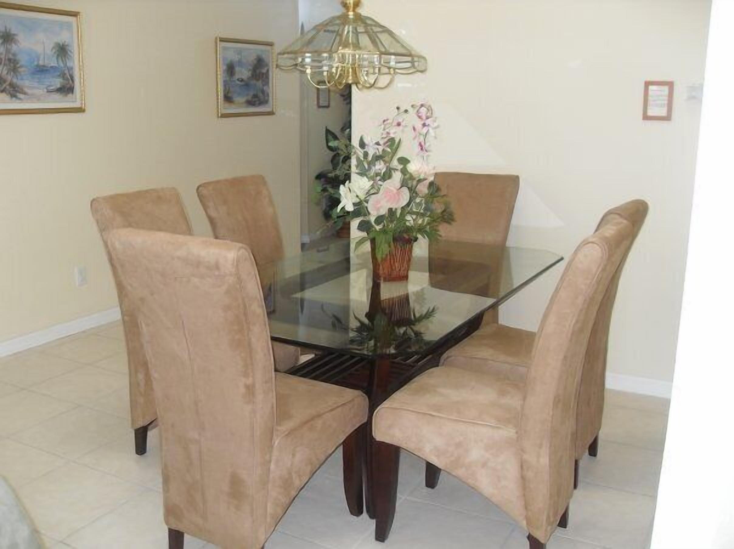 A dining table with a glass top surrounded by six beige chairs, a chandelier, and a vase with flowers.