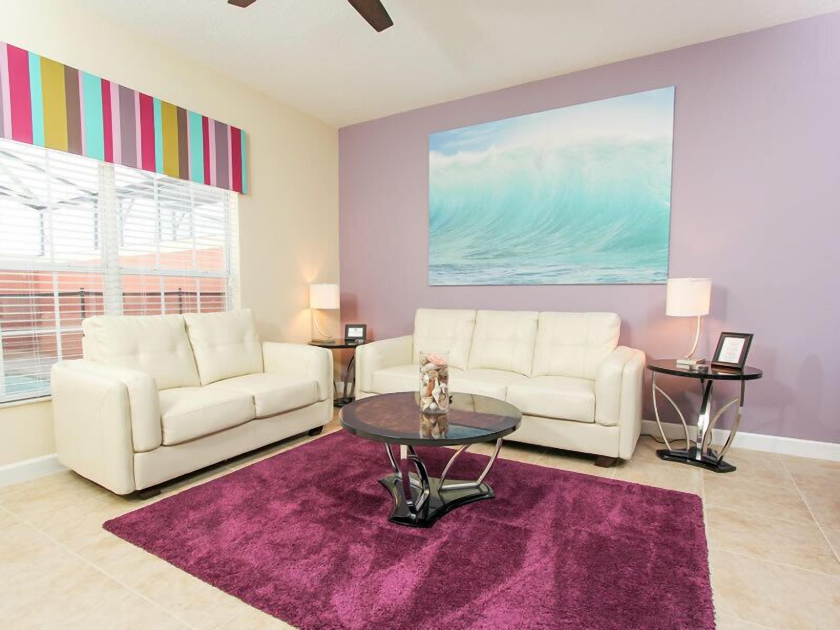 A living room with two white sofas, a round coffee table, a purple rug, a large painting of a wave, and a colorful striped wall.
