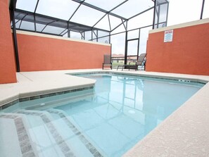 Una piscina interior con paredes de vidrio transparente, suelo de baldosas y algunas sillas de descanso.