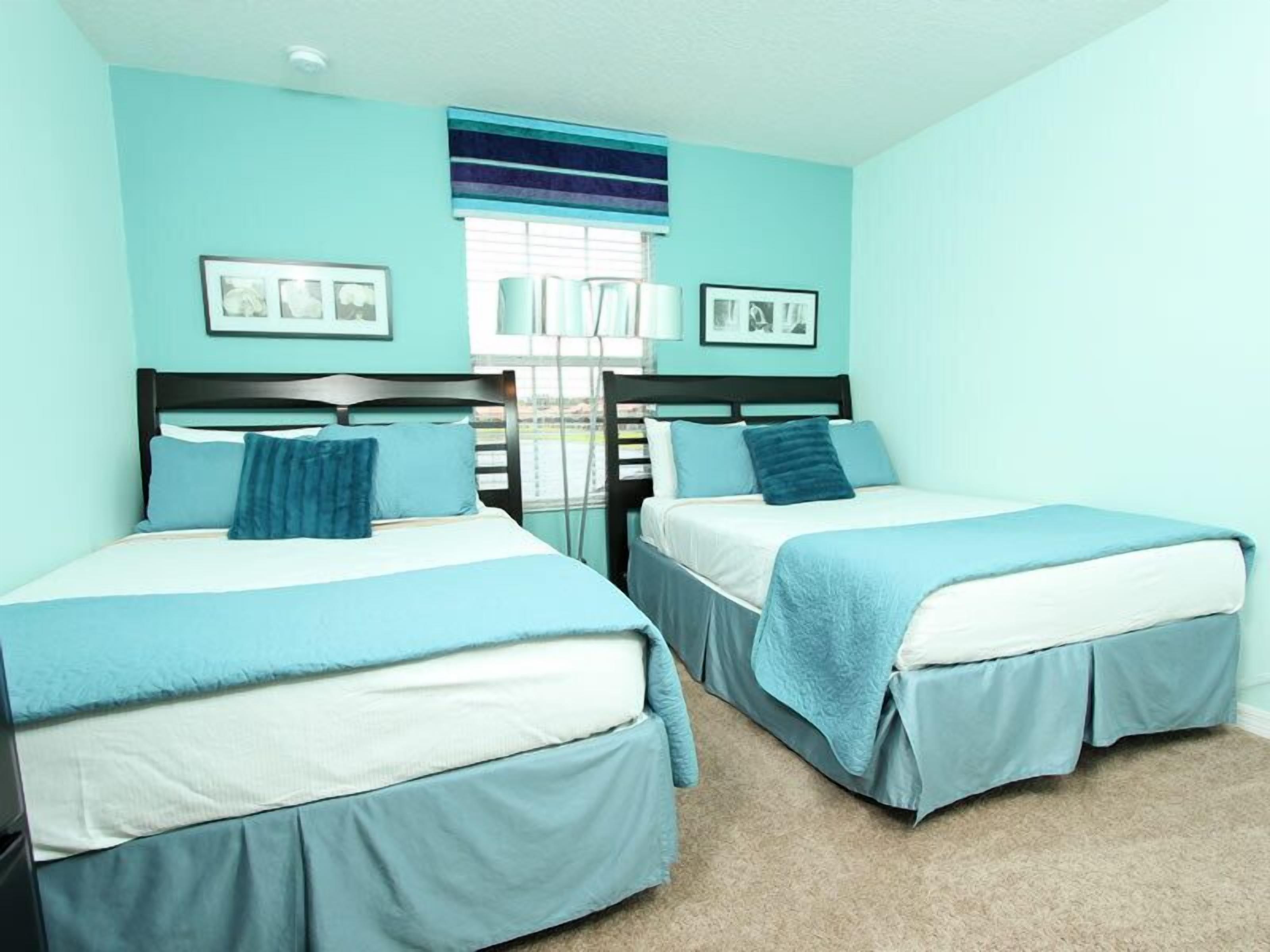 A room with two beds, each with a blue bedspread and matching pillows. There are framed pictures on the wall and a window with blinds.