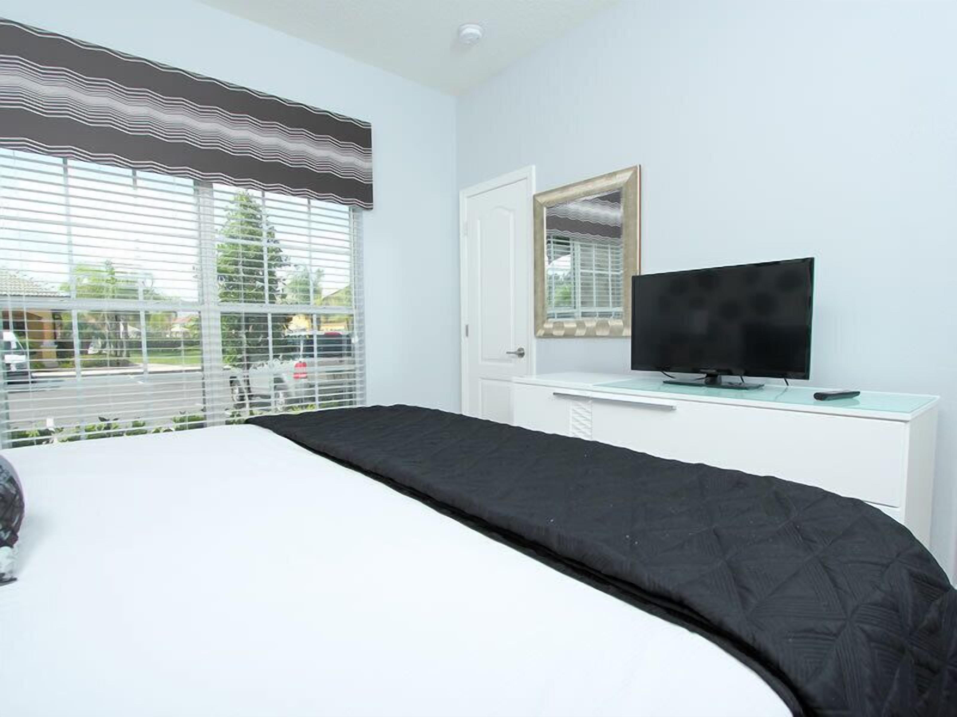 A bedroom with a bed, a TV on a white dresser, and a window with blinds.