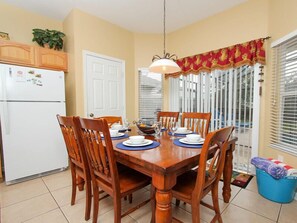 A dining area with a wooden table set for four, surrounded by chairs. There is a white refrigerator to the left, a door in the background, and a window with blinds to the right. - Ov2921 - Windsor Hills Resort - 6 Bed 4 Baths Villa (Kissimmee)