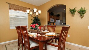 Comedor con una mesa de madera dispuesta para cuatro personas, rodeada de sillas, una araña y un ventanal con persianas.