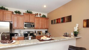 A modern kitchen with wooden cabinets, stainless steel appliances, and a marble countertop. There are three bar stools and a potted plant on the counter.