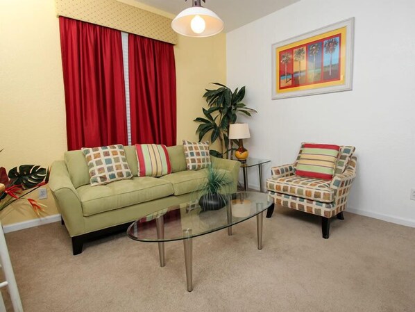 A living room with a green sofa, a striped armchair, a glass coffee table, a red curtain, a potted plant, and a framed painting on the wall.