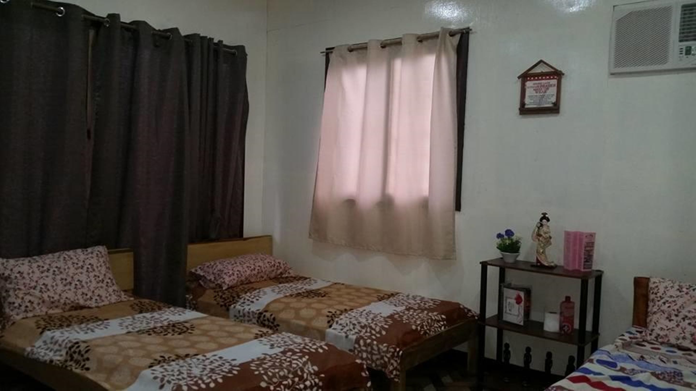 A room with two beds, a window with curtains, a small table with a plant, and a wall-mounted air conditioner.