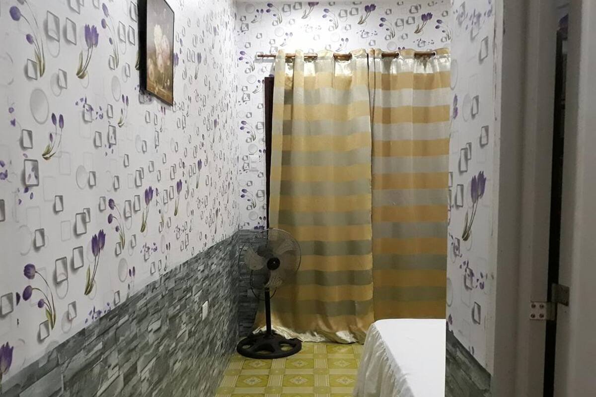 Une petite pièce étroite, avec un lit, un ventilateur sur pied et un rideau de douche.