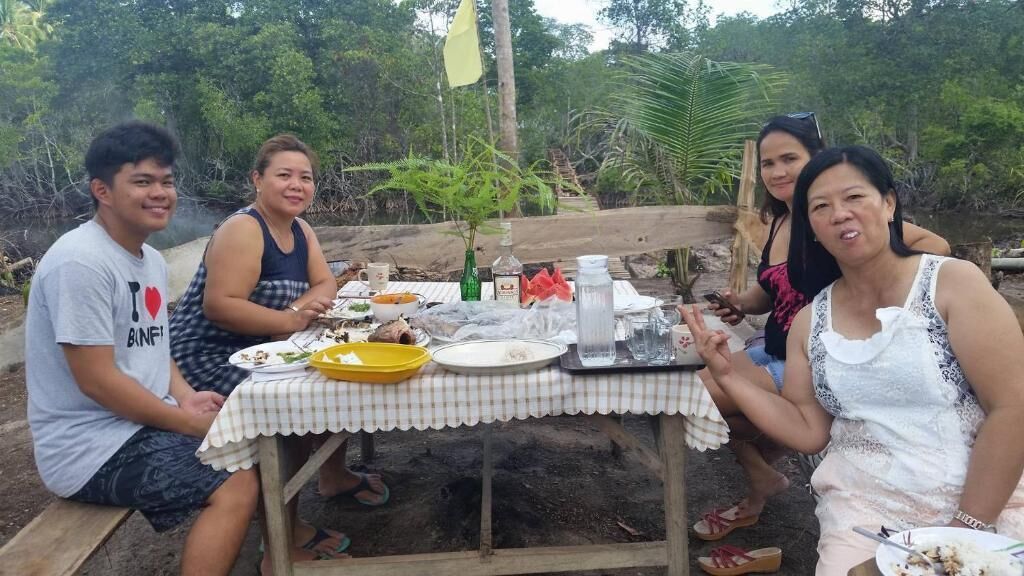 Un grupo de personas sentadas alrededor de una mesa con comida y bebidas, al aire libre con árboles de fondo.