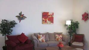 Uma sala de estar com um sofá vermelho, um sofá bege, uma mesa de centro de madeira e uma poltrona de vime. Há duas luminárias fixadas na parede e um quadro com flores.