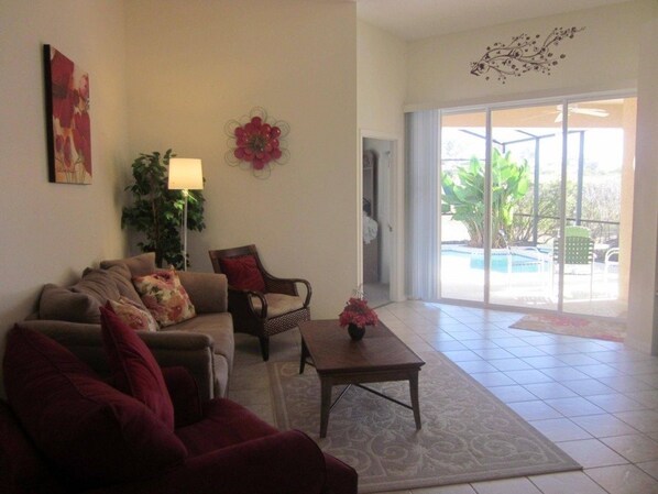 A living room with a sofa, armchair, and coffee table, a plant, and a view of the outside through a glass door.