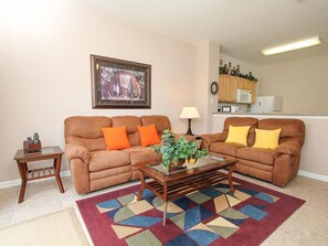 Un salon avec un canapé marron, deux coussins orange, une table basse en verre et un tapis coloré.