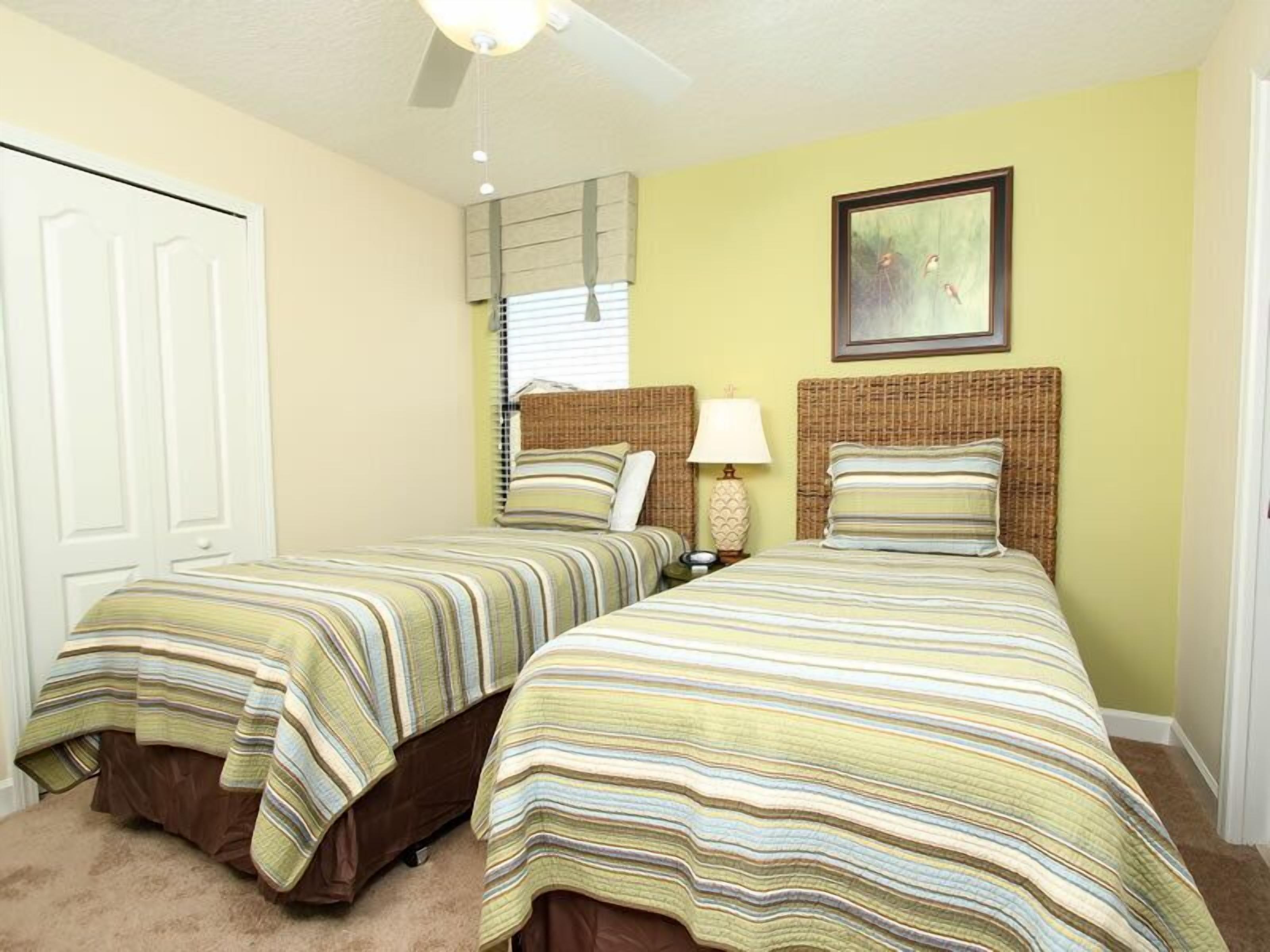 A room with two beds, each with a striped bedspread and a headboard. There is a window with blinds, a lamp on the nightstand, and a framed picture on the wall.