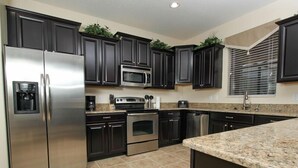 A modern kitchen with dark cabinetry, stainless steel appliances, a granite countertop, and a window with blinds. - Ov2856 - Champions Gate Resort - 6 Bed 6 Baths Villa (Davenport)