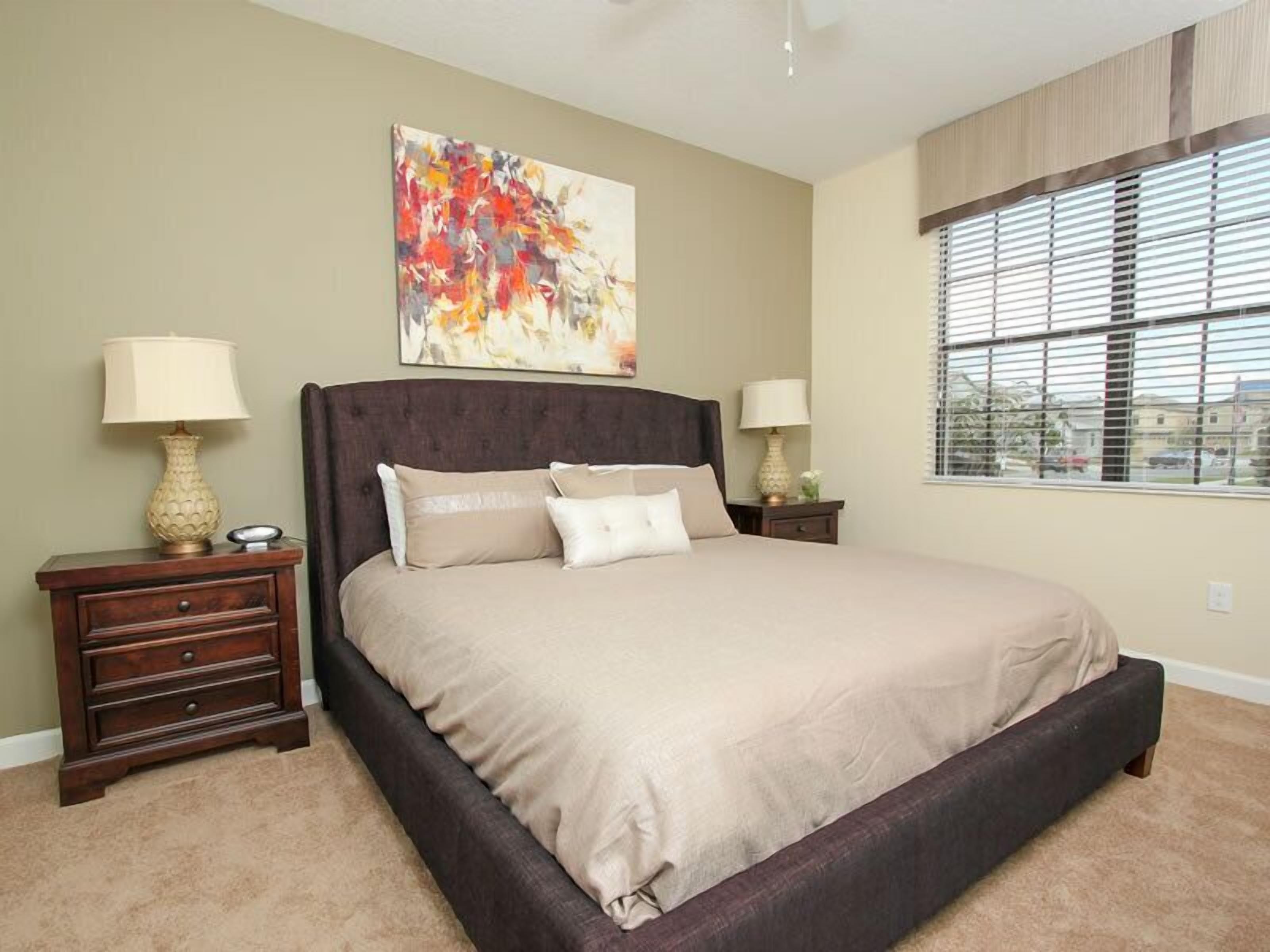 A bedroom with a large bed, two nightstands, a window with blinds, and a colorful abstract painting on the wall.