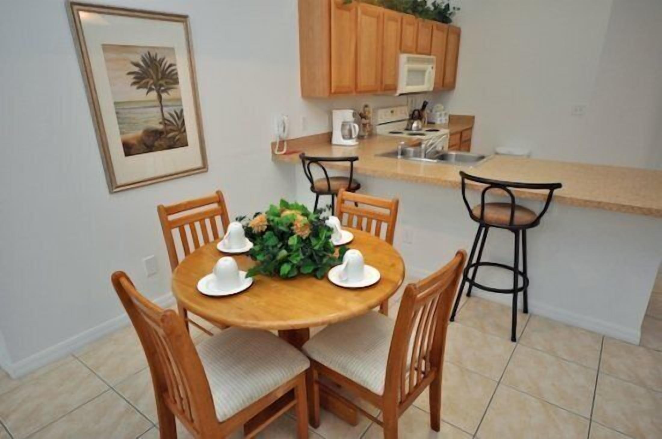 A dining area with a round wooden table set for six, surrounded by wooden chairs with cushioned seats. A small bar stool is positioned at the end of the counter.