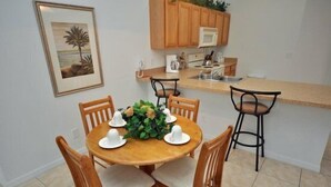 A dining area with a round wooden table set for six, surrounded by wooden chairs with cushioned seats. A small bar stool is positioned at the end of the counter.