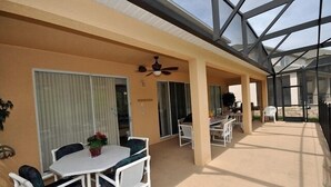 Un espace extérieur abrité, équipé d’une table et de chaises, d’un ventilateur de plafond et d’une paroi vitrée.