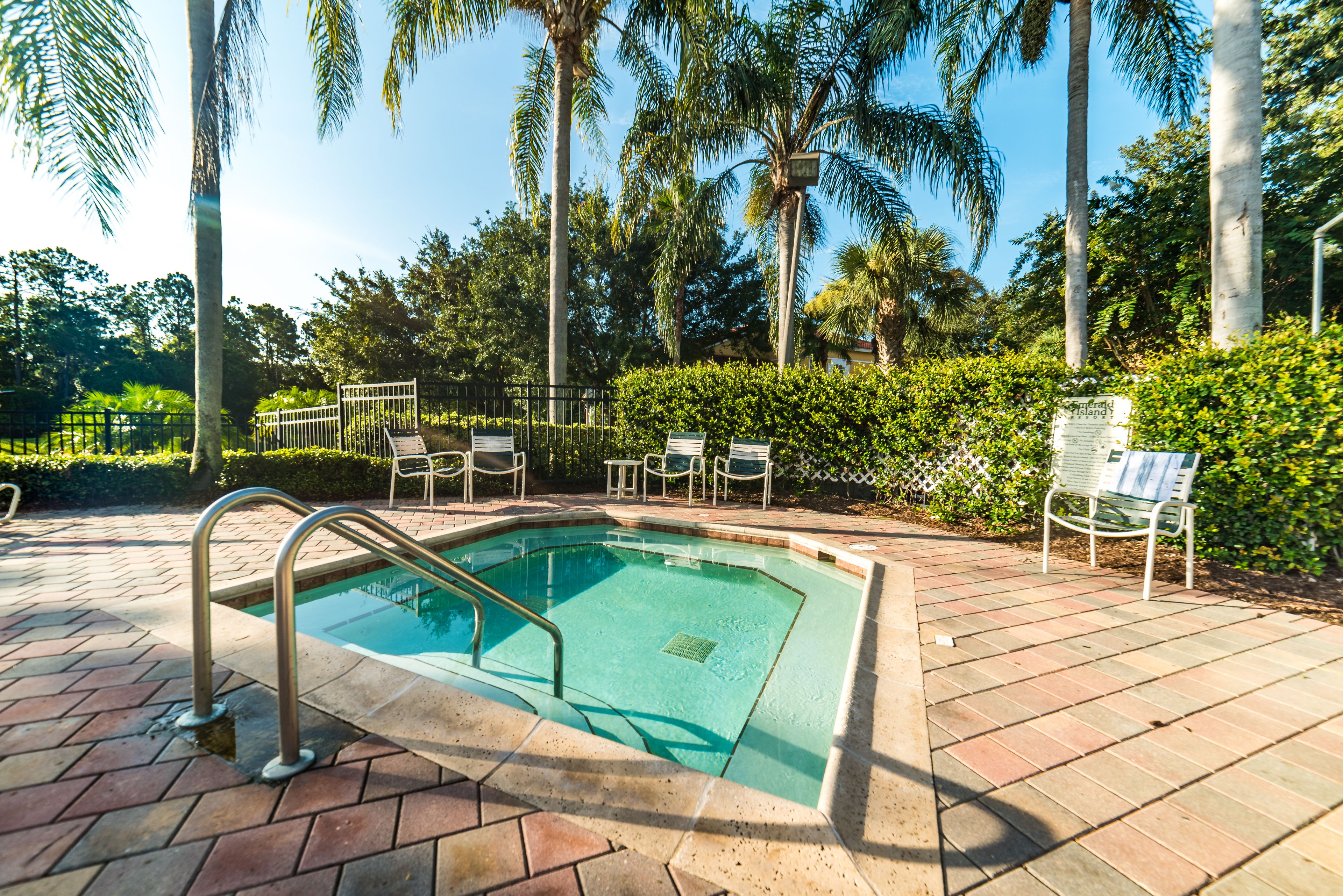 A swimming pool with a metal ladder, surrounded by a brick patio, palm trees, and outdoor seating.