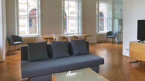 A modern living room with a grey sofa, a coffee table, and a dining area with a table and chairs. Large windows provide natural light and views of the outside. - Hofgärtnerhaus (Dresden)