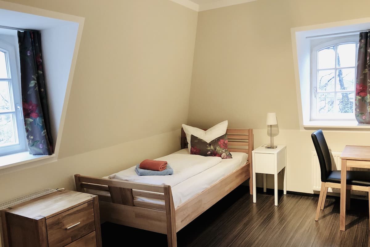 A small, well-lit room with a single bed, a desk, and a chair. There is a window with curtains, a radiator, and a wooden dresser.