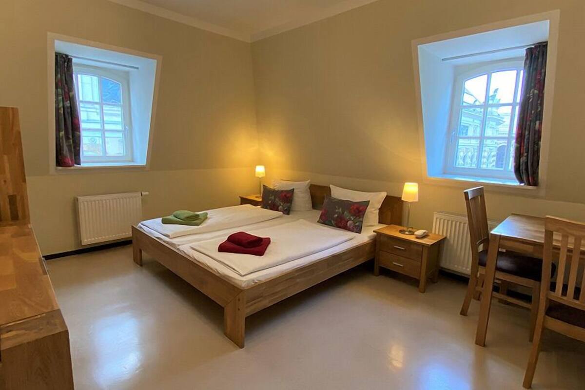 A bedroom with a wooden bed, two bedside tables with lamps, a chair, and two windows with curtains.