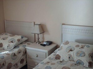 A bedroom with two beds, each with a patterned bedspread, a bedside table with a lamp, and a drawer.
