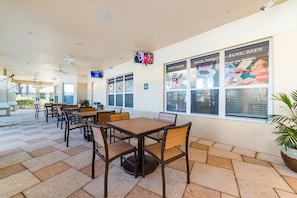 Guests can dine or relax in this outdoor seating area with tables and chairs, surrounded by potted plants and a TV.