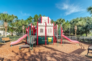 Children can play and explore in this castle-like play area with two slides, surrounded by a fence and benches for seating.