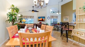 A dining area with a wooden table set for four, surrounded by wooden chairs. There is a ceiling fan above the table, and a chandelier hanging from the ceiling.