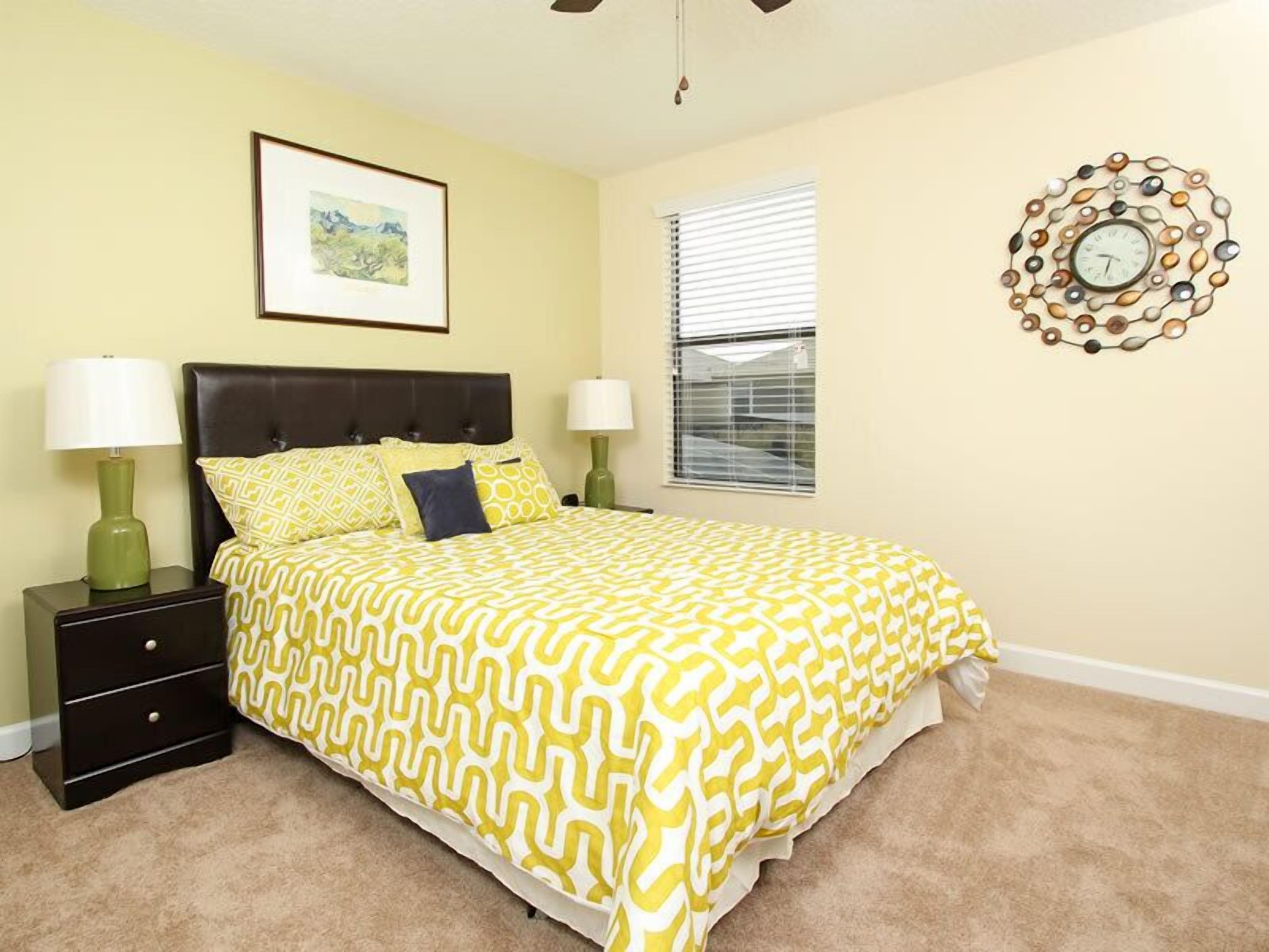 A bedroom with a bed, two bedside tables, a window with blinds, a framed picture on the wall, and a decorative clock.