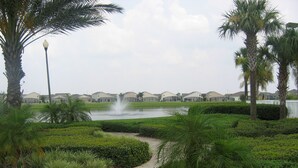 A landscaped area with a fountain, palm trees, and a pathway leading to a row of houses. - Ov2487 - Hampton Lakes - 5 Bed 3 Baths Villa (Davenport)