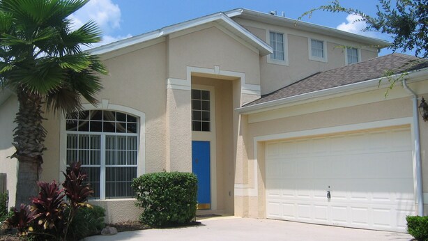 A two-story house with a garage, a blue door, and a palm tree in the front yard. - Ov2487 - Hampton Lakes - 5 Bed 3 Baths Villa (Davenport)