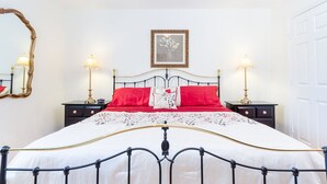 Cama bem arrumada com colcha branca e vermelha, com duas mesas de cabeceira com abajures, um quadro emoldurado acima da cama e um espelho na parede.