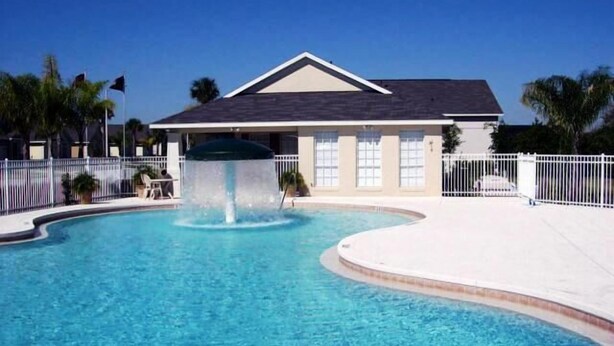 A swimming pool with a fountain, surrounded by a white fence, and a building with a dark roof in the background. - Ov1584 - Glenbrook Resort - 4 Bed 3 Baths Villa (Clermont)
