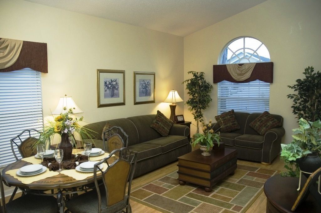 A living room with a sofa, two armchairs, a coffee table, and a dining table set for four. There are two lamps, a potted plant, and a window with blinds.