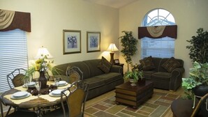A living room with a sofa, two armchairs, a coffee table, and a dining table set for four. There are two lamps, a potted plant, and a window with blinds. - Ov1584 - Glenbrook Resort - 4 Bed 3 Baths Villa (Clermont)