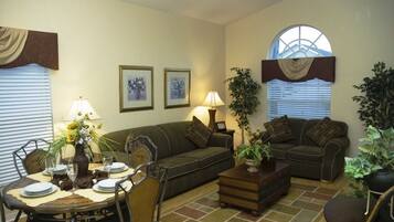 A living room with a sofa, two armchairs, a coffee table, and a dining table set for four. There are two lamps, a potted plant, and a window with blinds.