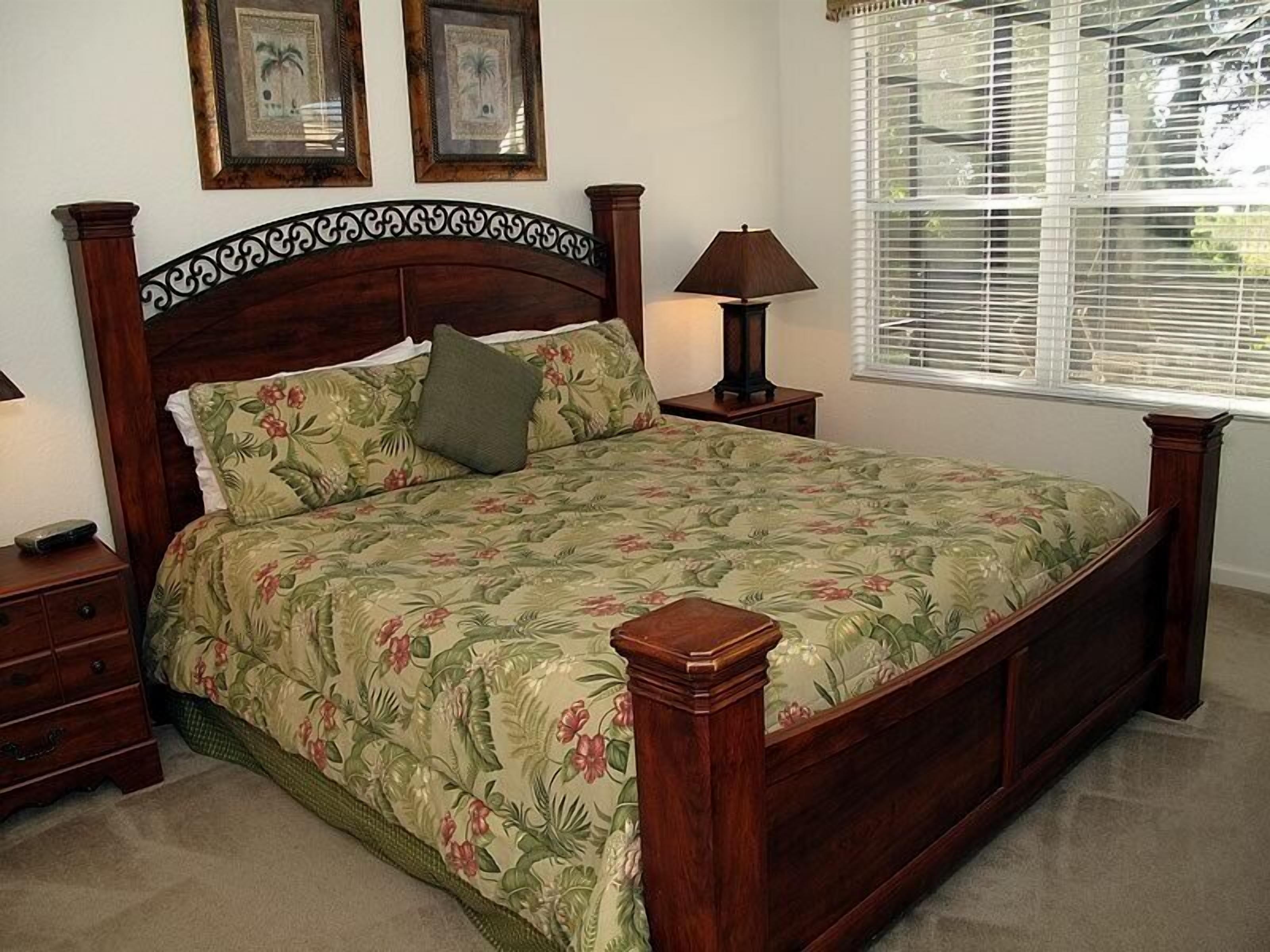 A bedroom with a wooden bed frame, floral bedding, a nightstand with a lamp, and a window with blinds.