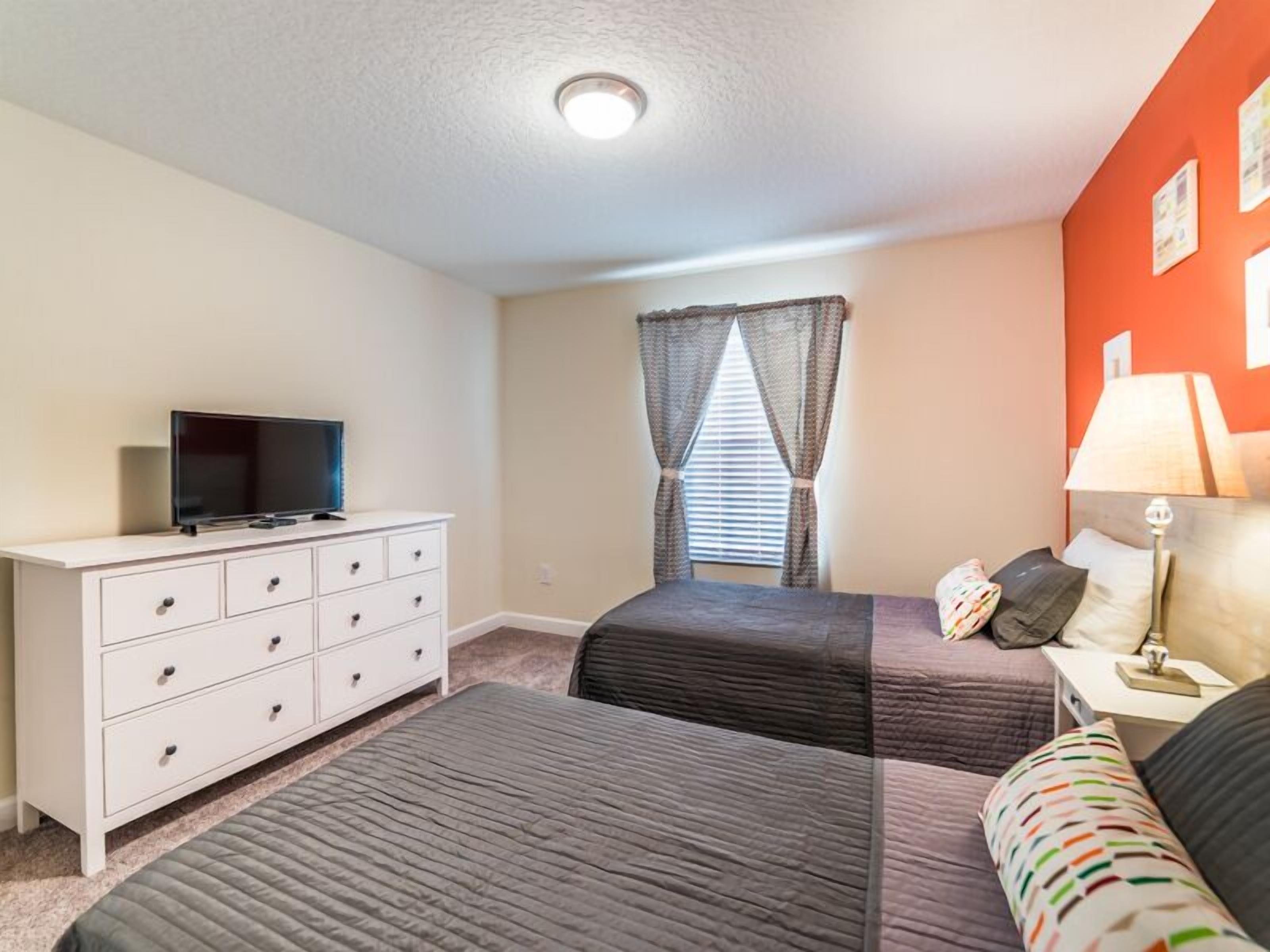 A bedroom with two beds, a TV on a dresser, a window with curtains, and a lamp on a bedside table.