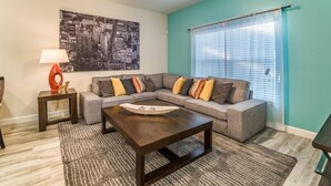 A living room with a sectional sofa, a coffee table, a rug, a lamp, and a cityscape painting on the wall. - Ov2069 - Paradise Palms - 2 Bed 1 Baths Villa (Kissimmee)